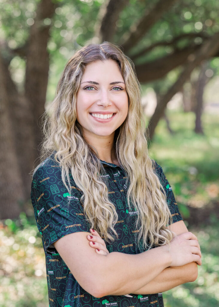 Hadley Green smiles at the camera while crossing her arms in front of green foliage