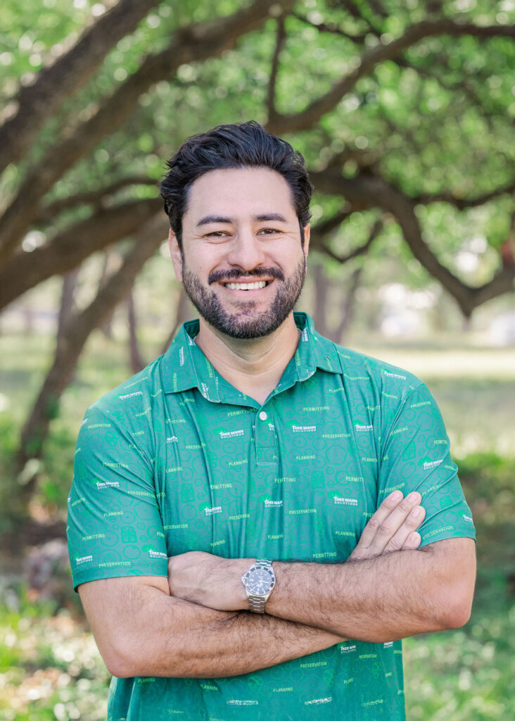 Jim Avila smiles at the camera wearing a green Tree Mann Solutions shirt with trees in the background